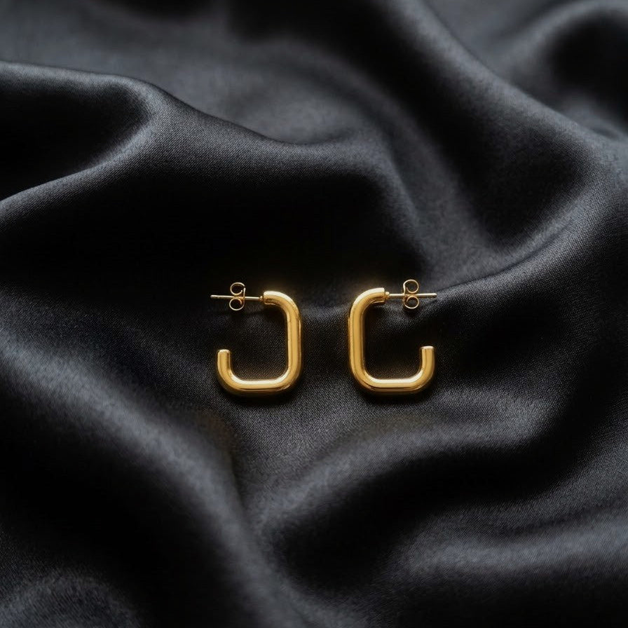 Pair of minimalist gold rectangular C-hoop earrings with stud backings resting on draped black satin fabric.