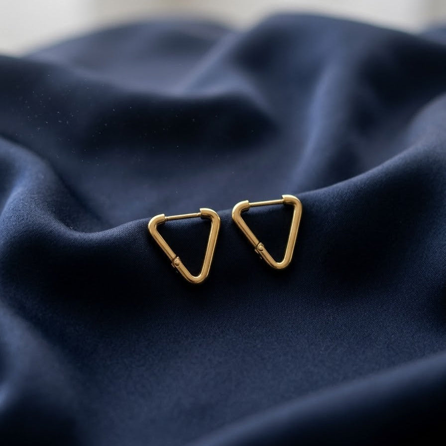 Pair of minimalist gold triangle-shaped huggie hoop earrings with a hinge closure resting on draped navy blue silk fabric