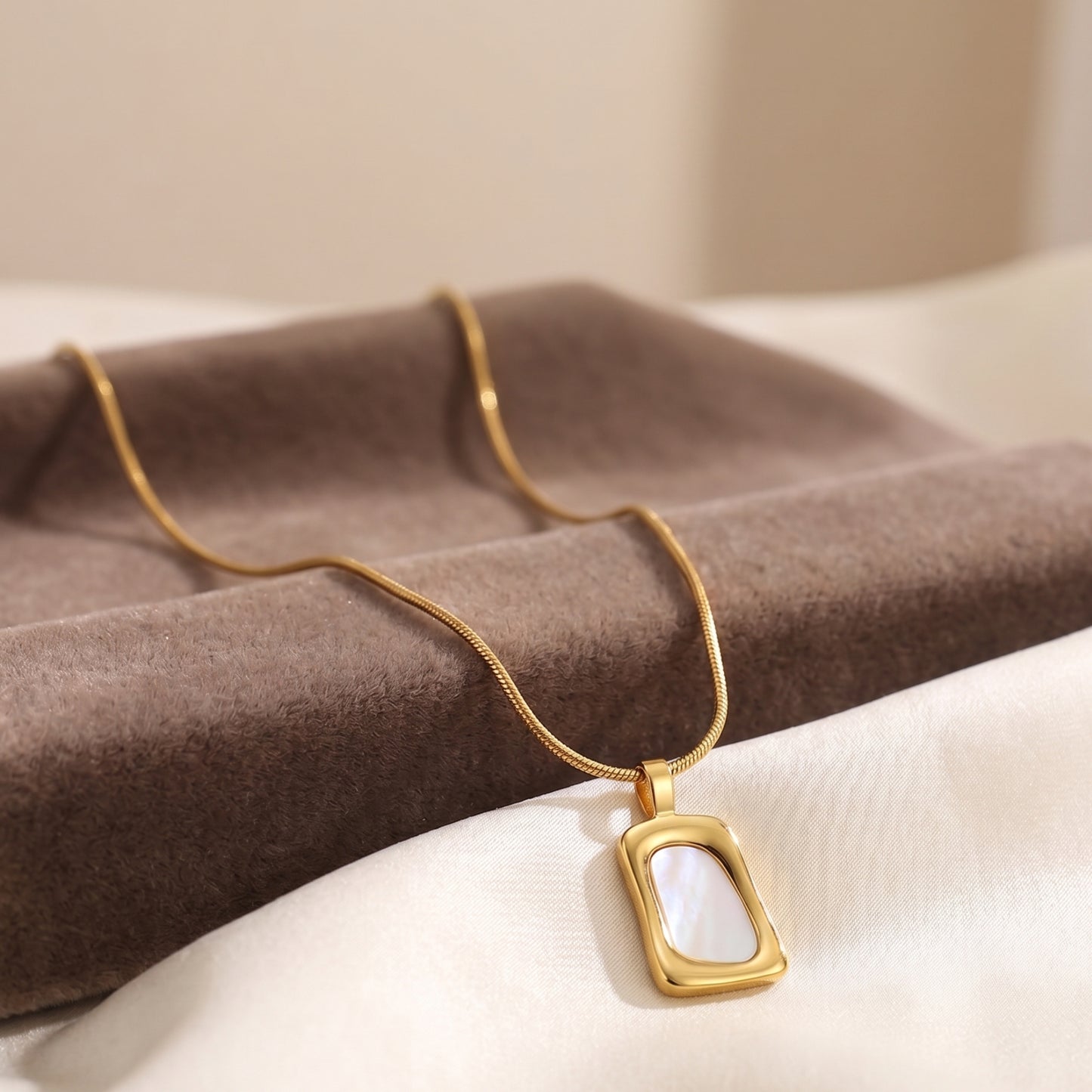Close-up of a gold geometric pendant necklace with white mother of pearl inlay on a beige jewelry display box.