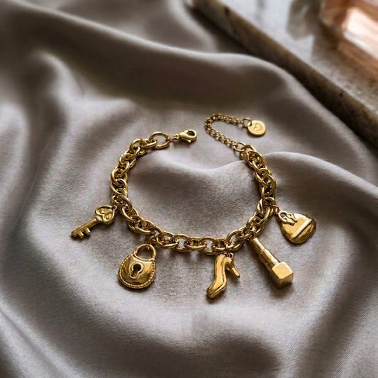 Gold-plated link chain charm bracelet featuring a lipstick, high heel shoe, handbag, key, and padlock charms on grey silk fabric.