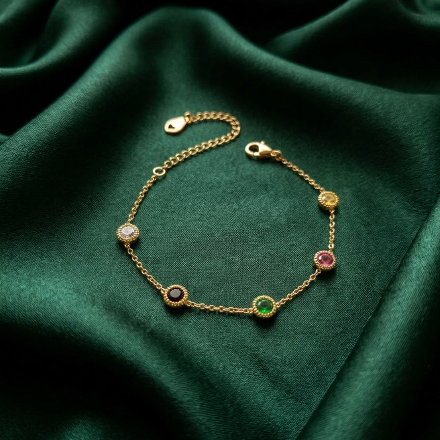 Dainty gold-plated station bracelet featuring five round multi-colored stones in bezel settings, displayed on emerald green silk fabric.