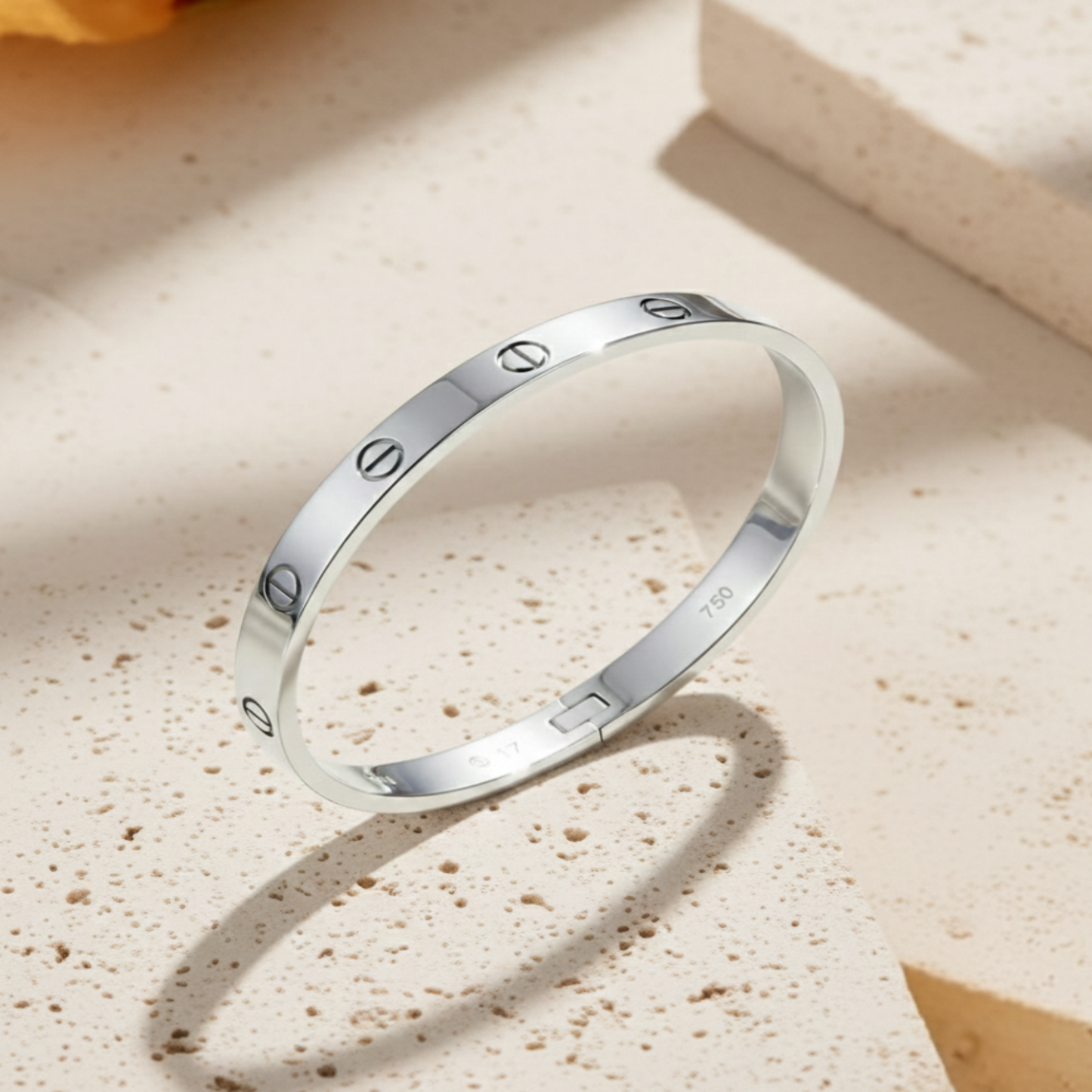 A polished silver screw motif bangle bracelet made of rhodium-plated stainless steel, displayed on a neutral cream surface next to a decorative ceramic bowl.