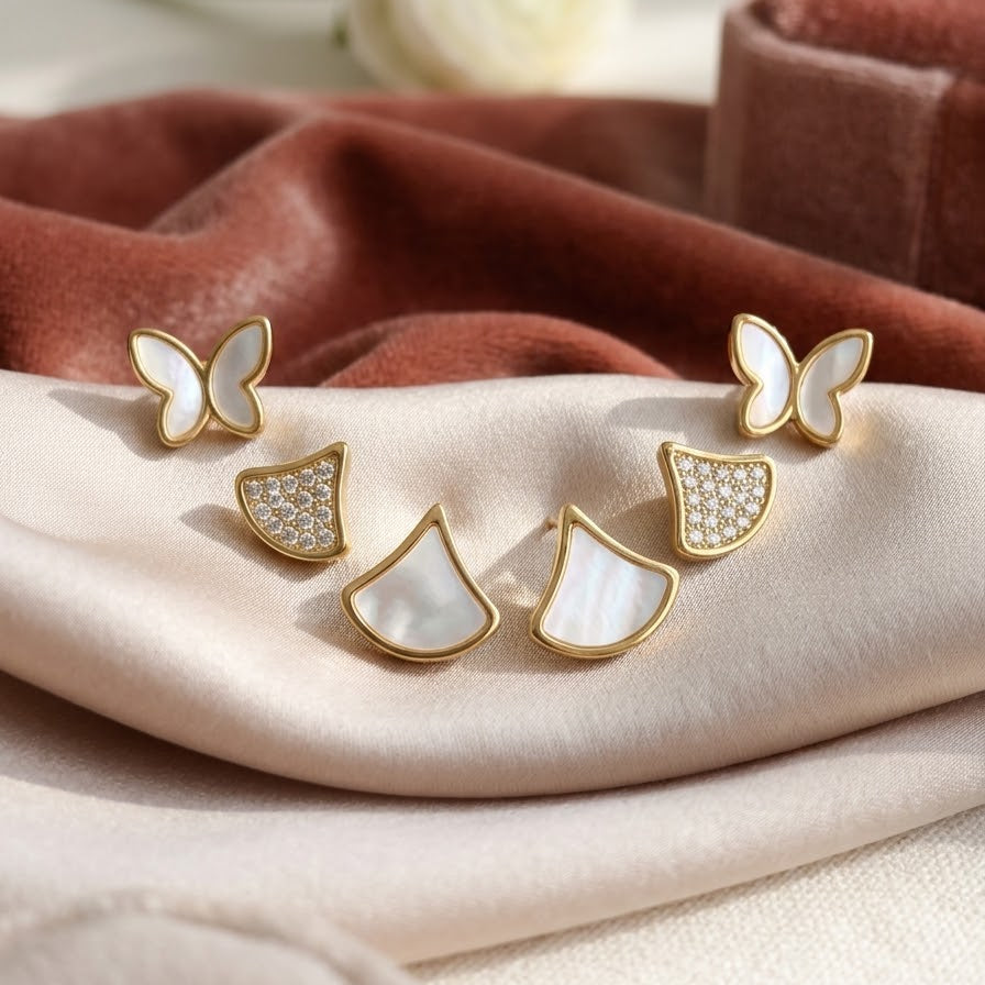 Curated set of gold-plated stud earrings featuring Mother of Pearl butterflies and fan shapes alongside crystal pavé fan studs on pink velvet.