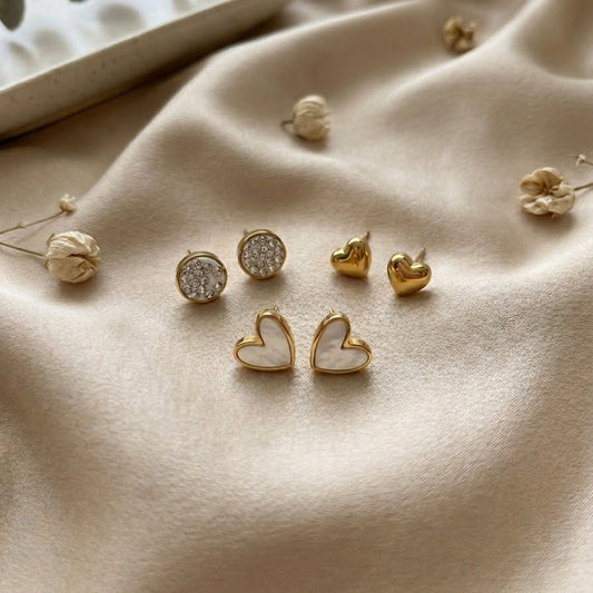 Collection of gold-plated stud earrings featuring white Mother of Pearl hearts, crystal-encrusted circles, and puffed gold hearts on cream silk.