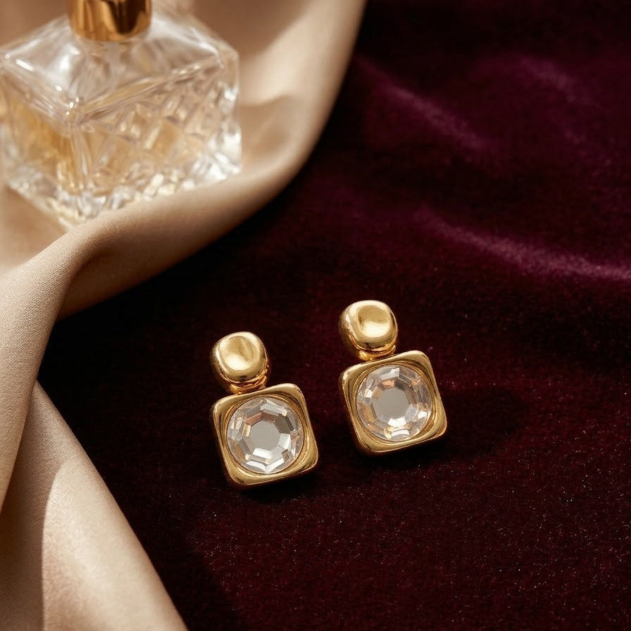 Pair of square gold-plated stud earrings with large central faceted crystals resting on dark burgundy velvet next to a perfume bottle.