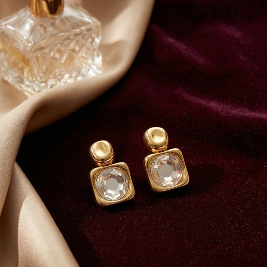 Pair of square gold-plated stud earrings with large central faceted crystals resting on dark burgundy velvet next to a perfume bottle.