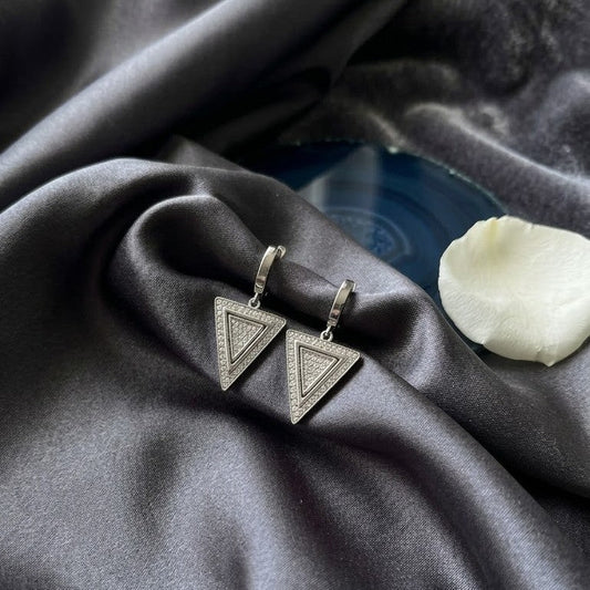 Pair of silver triangle-shaped drop earrings with pavé crystal inlay resting on draped dark grey satin fabric.