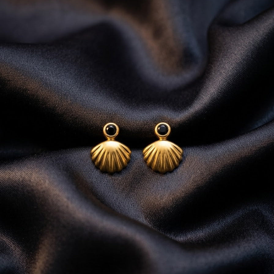 Pair of minimalist gold scallop shell stud earrings with small black stones centered on dark grey draped silk fabric.