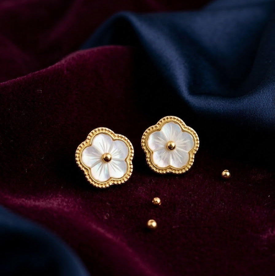 Pair of gold-plated flower stud earrings with iridescent white petals and a beaded gold rim, placed on dark purple and navy blue velvet fabric.