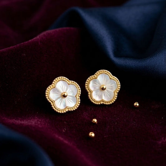Pair of gold-plated flower stud earrings with iridescent white petals and a beaded gold rim, placed on dark purple and navy blue velvet fabric.