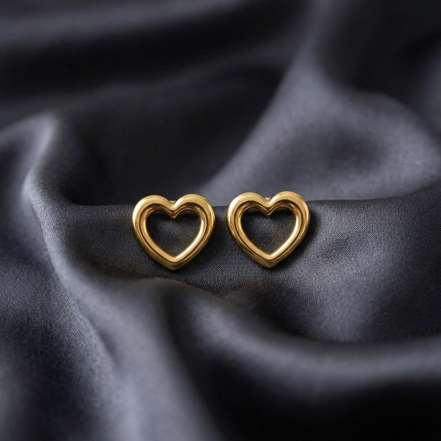 Pair of minimalist chunky gold heart-shaped stud earrings with an open center, resting on soft, draped charcoal grey silk fabric.