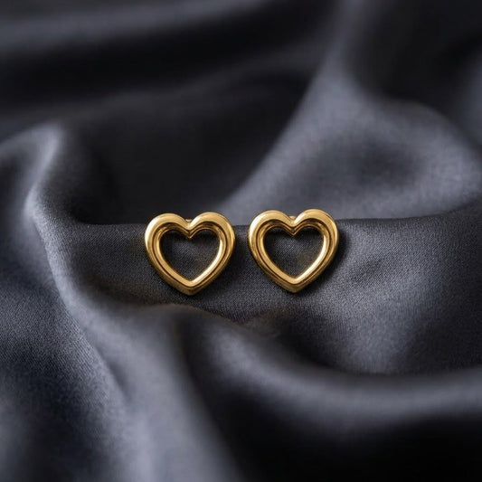 Pair of minimalist chunky gold heart-shaped stud earrings with an open center, resting on soft, draped charcoal grey silk fabric.