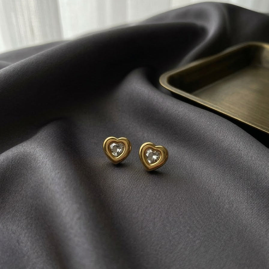 Pair of gold-plated heart-shaped stud earrings with a central clear crystal inlay, resting on dark charcoal grey satin fabric.