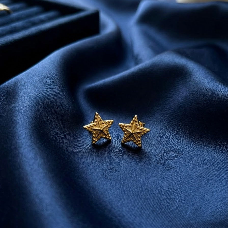 Pair of textured gold five-point star stud earrings with beaded detailing, resting on draped navy blue satin fabric