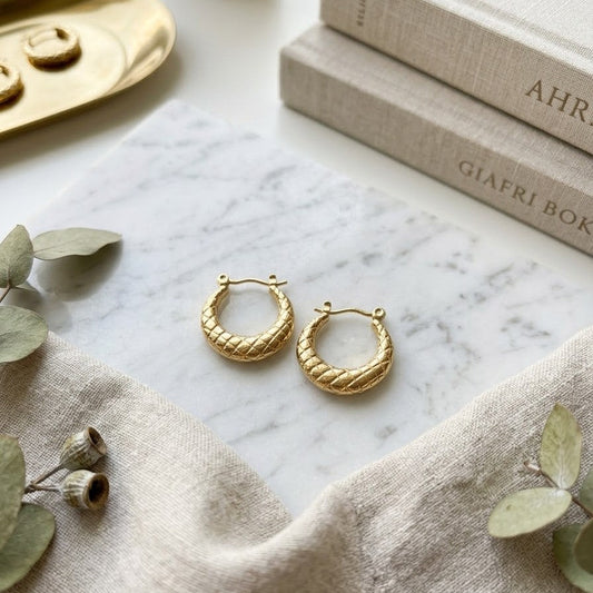 Pair of gold-plated hoop earrings with a quilted lattice texture resting on a white marble slab surrounded by linen books and dried eucalyptus.