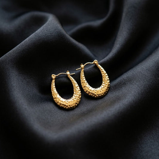 Pair of gold-plated oval hoop earrings with a hammered textured surface resting on draped black satin fabric.