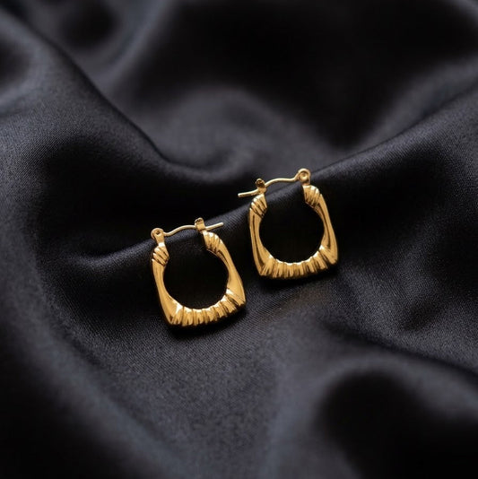 Pair of square gold-plated hoop earrings with a ribbed sunburst texture resting on draped black silk fabric.