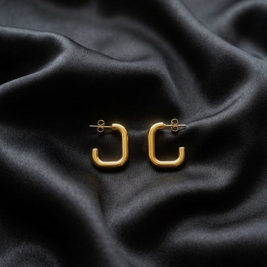Pair of minimalist gold rectangular C-hoop earrings with stud backings resting on draped black satin fabric.