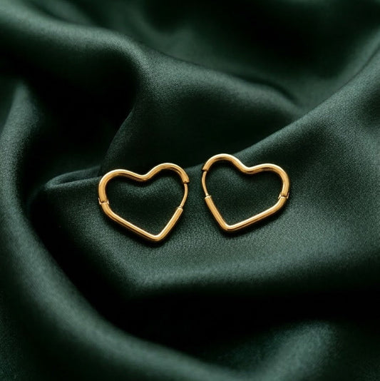 Pair of minimalist gold heart-shaped hoop earrings with a hinge closure resting on draped emerald green satin fabric
