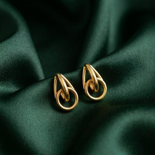 Pair of gold-plated geometric drop earrings with interlocking teardrop and circular links resting on draped emerald green satin fabric