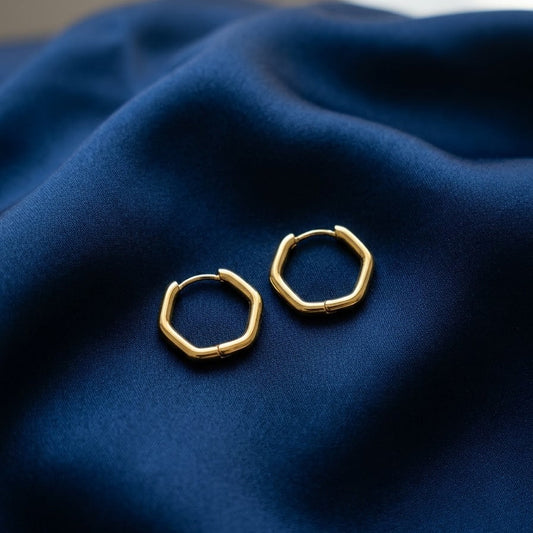 Pair of minimalist gold hexagon-shaped huggie hoop earrings with a hinge closure resting on draped royal blue silk fabric