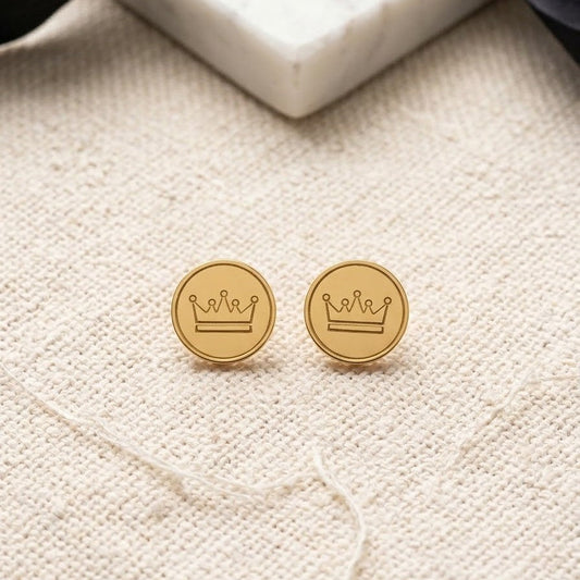 Pair of gold-plated circular stud earrings with an engraved crown design resting on a textured cream fabric background