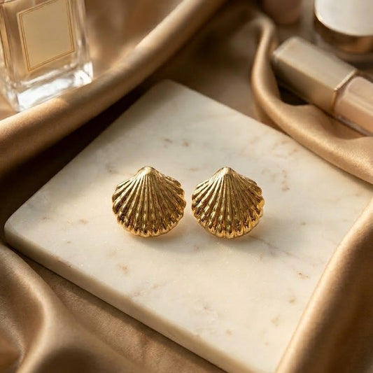 Pair of 3D textured gold scallop shell stud earrings resting on a white marble slab surrounded by gold silk fabric and perfume