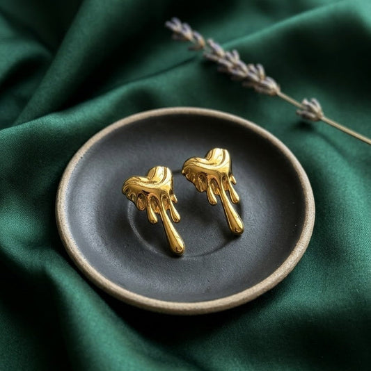 Pair of gold-plated stud earrings in a melting heart silhouette with a liquid drip effect, resting on a dark ceramic tray over emerald green fabric