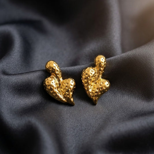 Pair of gold-plated abstract heart-shaped statement stud earrings with a hammered, dimpled texture resting on draped dark grey satin fabric