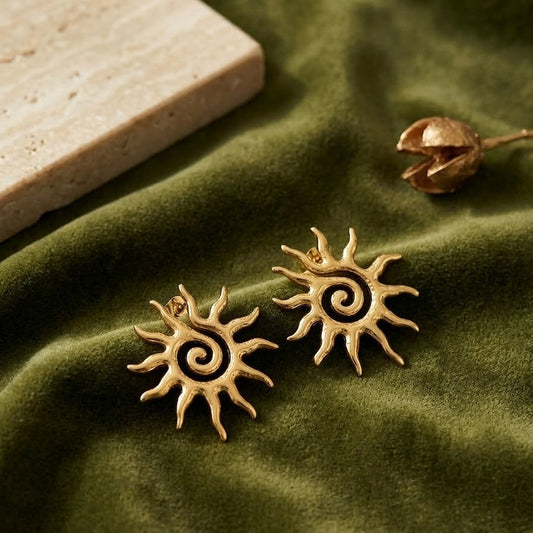 Pair of gold-plated celestial sun earrings with a spiral center and wavy sunburst rays resting on olive green velvet fabric near a marble slab