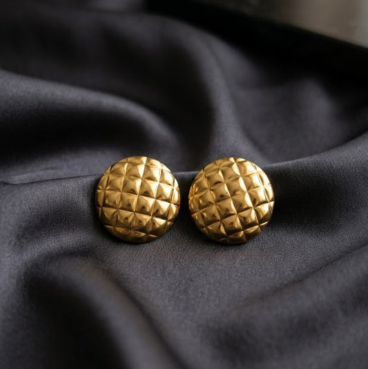 Pair of gold-plated round stud earrings with a 3D quilted diamond texture resting on draped dark grey satin fabric.