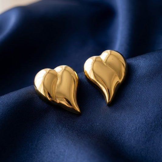 Pair of large gold-plated chunky puffy heart-shaped stud earrings with a high-shine mirror finish resting on draped navy blue silk fabric.