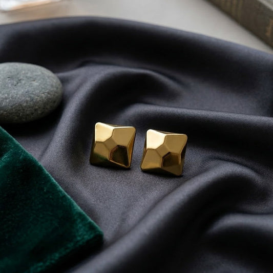 Pair of gold-plated 3D geometric pyramid-shaped stud earrings with a faceted mirror finish resting on draped charcoal grey silk fabric.