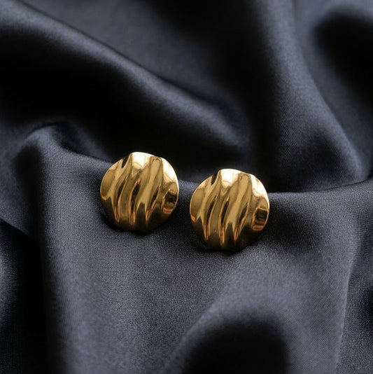 Pair of gold-plated round stud earrings with a wavy, rippled molten texture resting on draped black silk fabric
