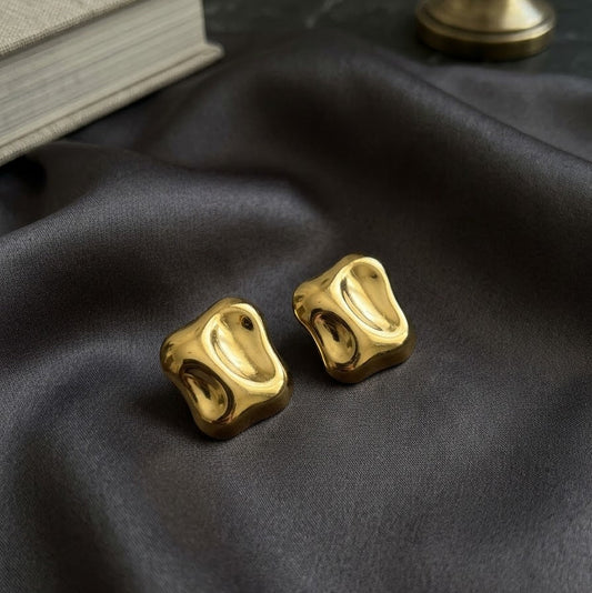 Pair of chunky gold-plated statement stud earrings with an organic molten square texture resting on draped dark grey satin fabric