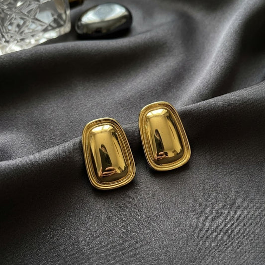 Pair of chunky gold-plated geometric statement stud earrings with a raised domed surface resting on draped charcoal grey silk fabric near a perfume bottle