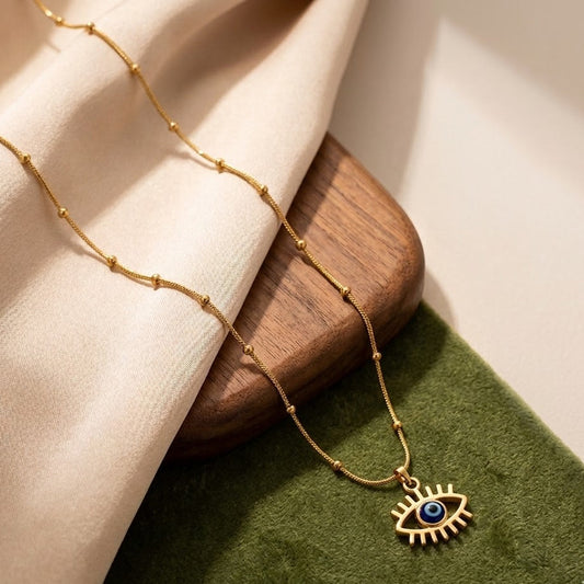 Gold-plated Evil Eye pendant necklace with blue eye charm on a satellite bead chain over a wooden and green velvet background.