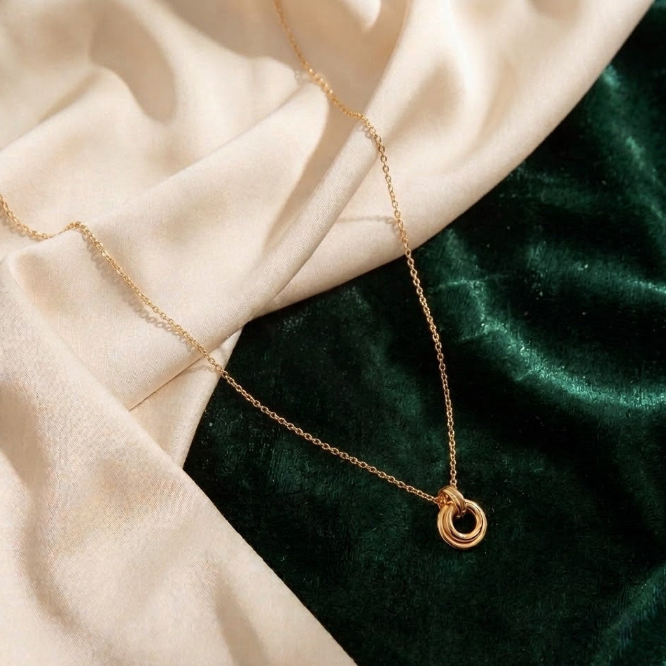 Dainty gold necklace with two interlocking circular rings on a fine link chain, draped over cream silk and emerald green velvet fabric.