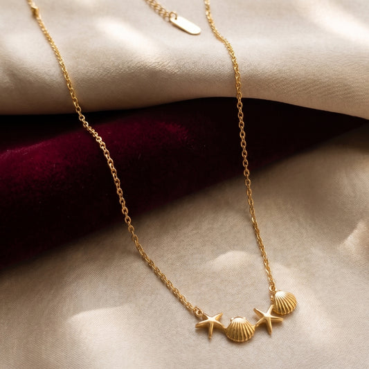 Dainty gold-plated necklace with three scallop shell and two starfish charms resting on beige silk and red velvet fabric.