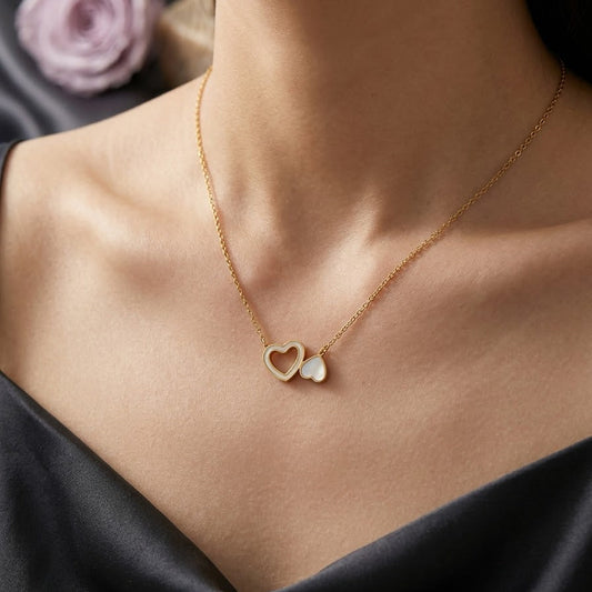 Model wearing a dainty gold necklace with two interlocking hearts—one open gold heart and one solid heart with a white pearl inlay.