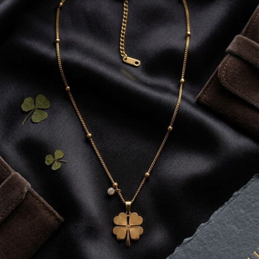 Gold-plated four-leaf clover pendant necklace with a small zircon crystal charm on a satellite bead chain, displayed on black silk fabric.