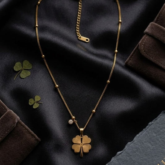 Gold-plated four-leaf clover pendant necklace with a small zircon crystal charm on a satellite bead chain, displayed on black silk fabric.