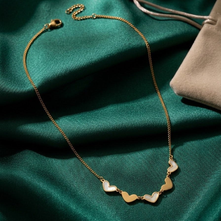 Dainty gold station necklace with five heart charms, featuring alternating gold and white pearl inlays, displayed on emerald green silk fabric.