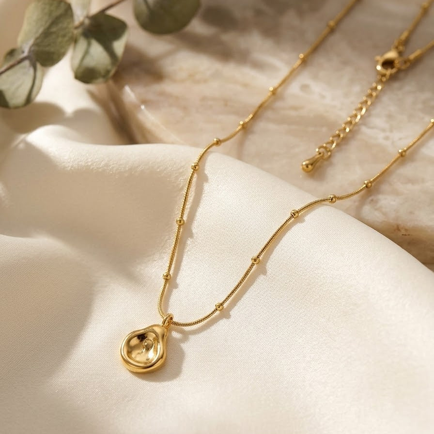 Dainty gold-plated necklace with an abstract molten teardrop pendant on a satellite bead chain, draped over white silk and a natural stone surface.