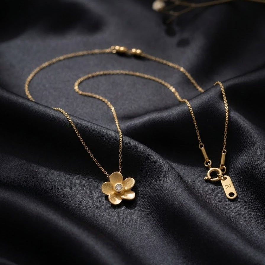 Dainty gold-plated flower necklace with brushed matte petals and a center zircon stone, displayed on elegant black silk and white satin backdrops.