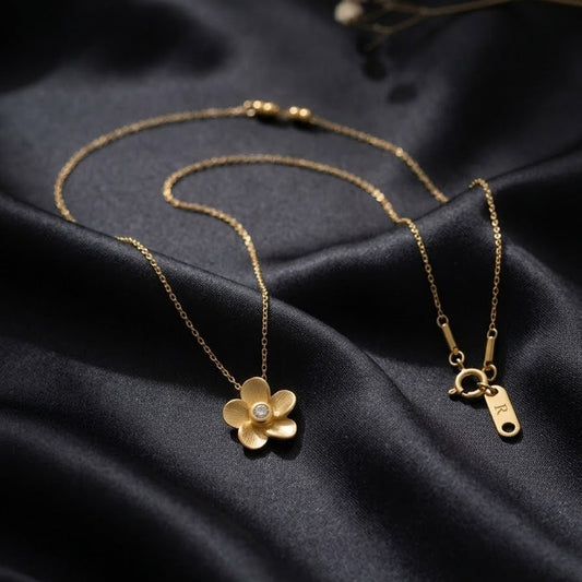Dainty gold-plated flower necklace with brushed matte petals and a center zircon stone, displayed on elegant black silk and white satin backdrops.