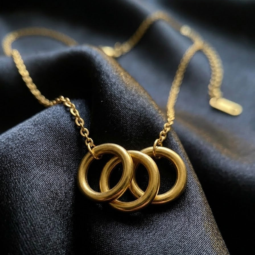 Dainty gold necklace with three interlocking circular rings on a fine link chain, displayed on elegant black silk fabric with a rose petal.