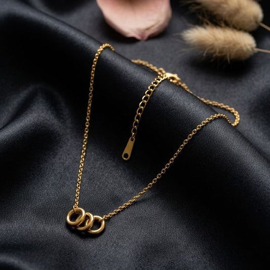 Dainty gold necklace with three interlocking circular rings on a fine link chain, displayed on elegant black silk fabric with a rose petal.
