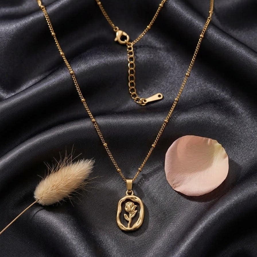 Gold-plated oval pendant necklace with a cut-out rose design on a satellite bead chain, displayed on black silk and beige velvet backgrounds with a rose petal.
