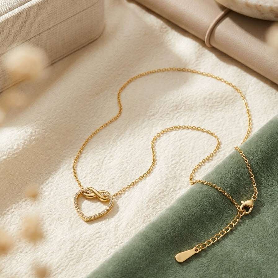 Dainty gold-plated necklace with an interlocking infinity loop and pavé crystal heart pendant, displayed on elegant black and champagne silk backdrops.